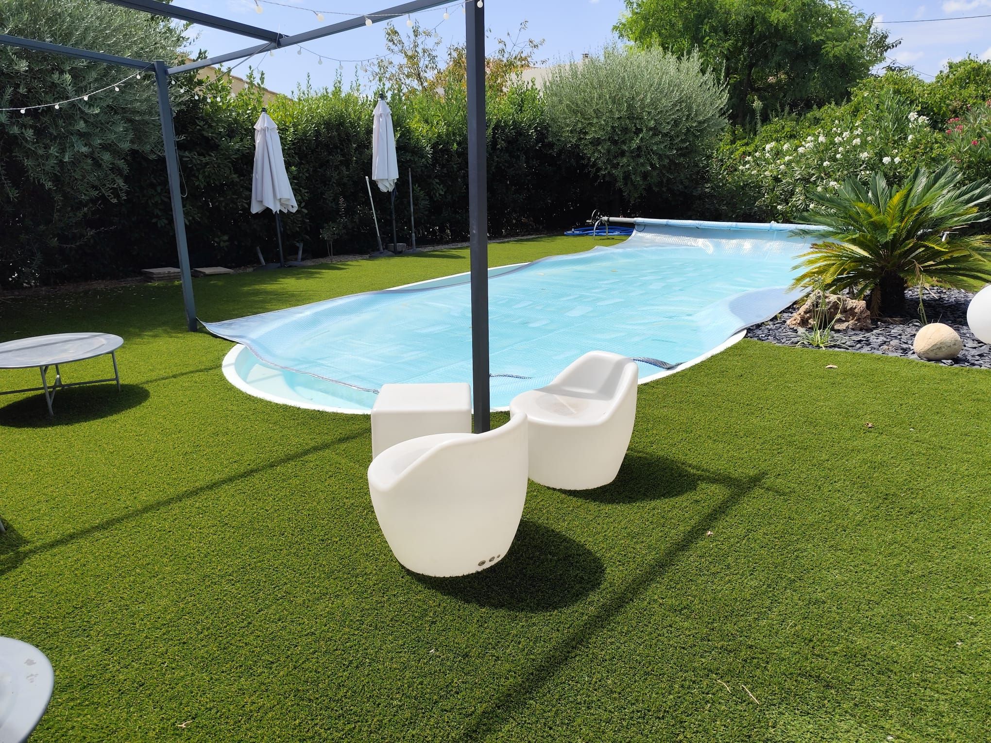 Piscine entourée de gazon synthétique, fauteuils blancs et voile d’ombrage